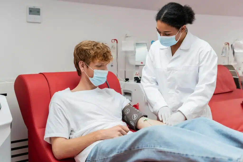 multiracial doctor in white coat and medical mask looking at redhead volunteer with blood pressure