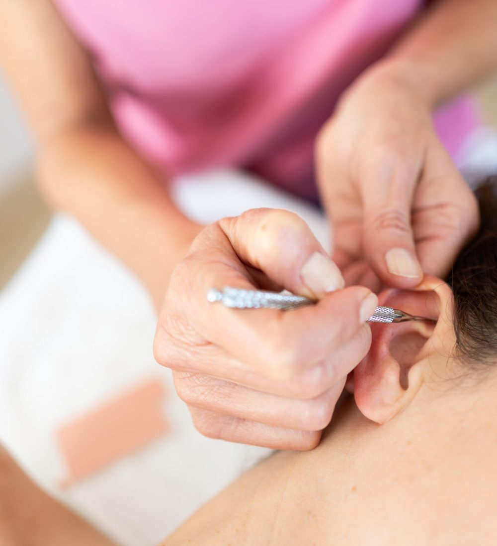 Crop professional beautician applying auriculotherapy ear acupuncture techniques of female client lying in modern beauty salon during facial treatment